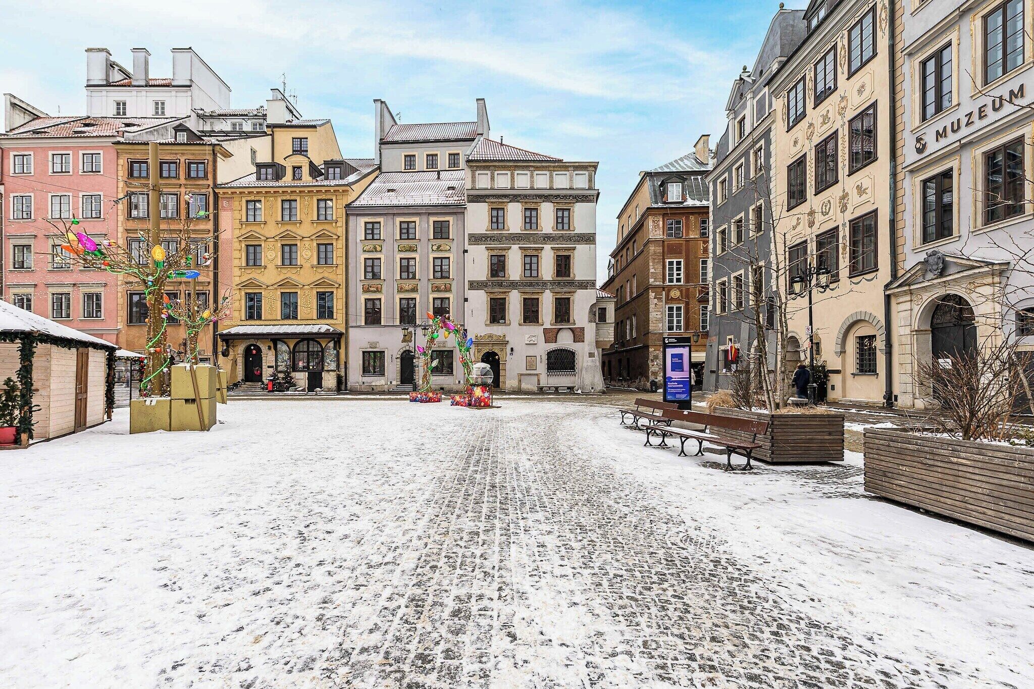 Een met sneeuw bedekte straat met kinderafsluitingen, historische gebouwen, banken en een bord voor een museum.