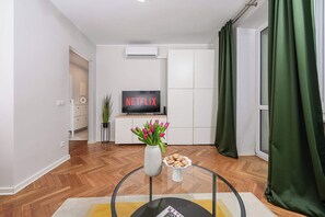 Ein modernes Wohnzimmer mit Flachbildfernseher, einer Blumenvase, einem Teller mit Keksen und einem Holzboden mit Fischgrätmuster.
