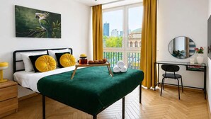 Habitación de hotel con cama, escritorio, silla, ventana con cortinas, un cuadro en la pared y vistas a la ciudad.