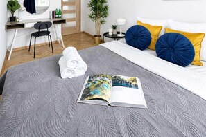 Ein ordentlich bezogenes Bett mit grauer, strukturierter Bettdecke, ein Paar aufgerollte Handtücher und eine offene Zeitschrift darauf.
