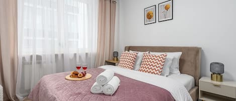 Habitació d'hotel amb un llit, una tauleta de nit, una làmpada i una finestra amb cortines translúcides.