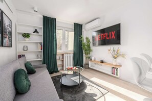 A modern living room with a grey sofa, a glass coffee table, a bookshelf, a TV mounted on the wall, and a window with curtains. - Modern Coastal Home with Scenic Terrace Views (Warszawa)
