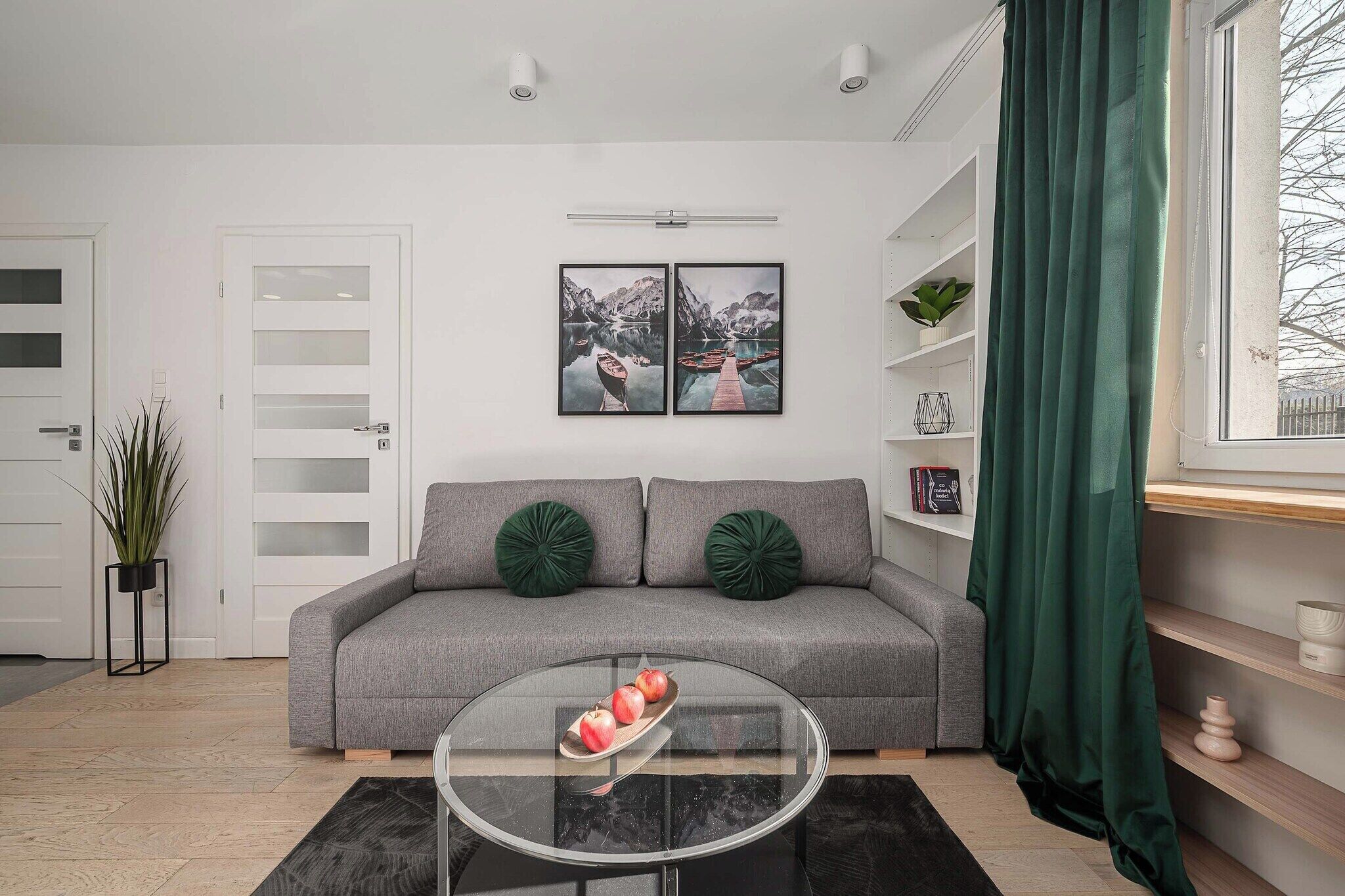 A living room with a grey sofa, two green cushions, a glass coffee table with apples, and a window with a green curtain.