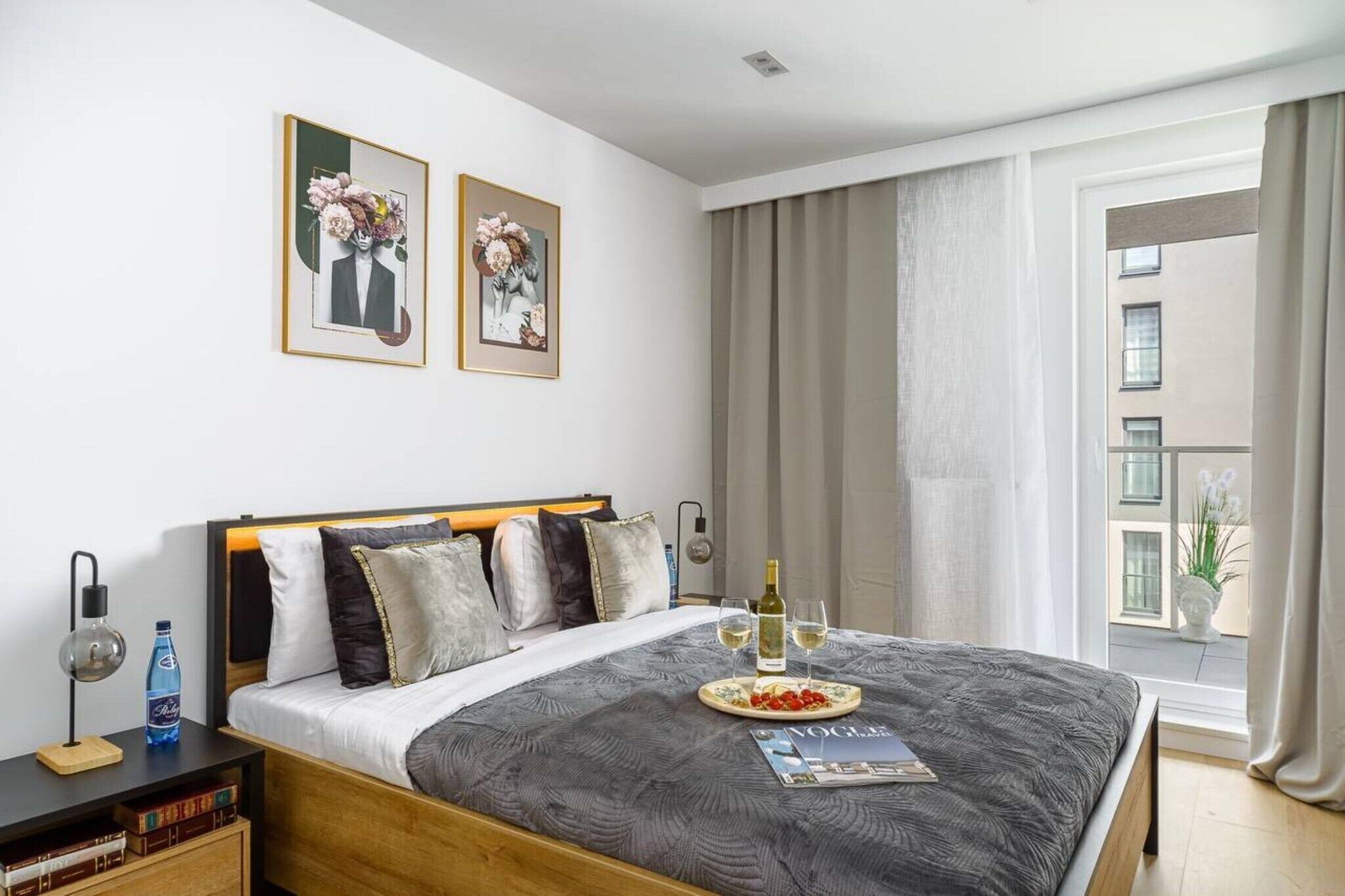 Una habitación de hotel con una cama grande, una mesita de noche con lámpara, una revista y una botella de vino.