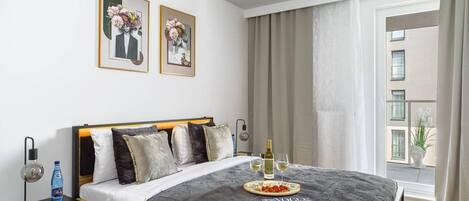 Una habitación de hotel con una cama grande, una mesita de noche con lámpara, una revista y una botella de vino.