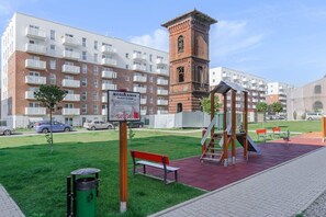 Ein Spielplatz mit Schaukeln und einer Bank, ein Backsteinturm und ein Hinweisschild in einem Wohngebiet.