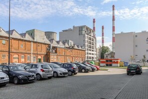 Ein Parkplatz mit mehreren Autos, ein Gebäude mit Backsteinfassade und Industriegebäuden mit Schornsteinen im Hintergrund.