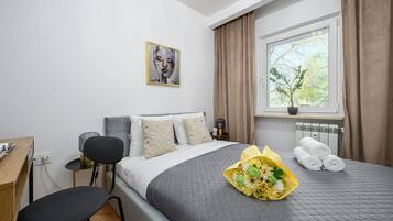 A hotel room with a bed, a desk, a chair, a window with curtains, and a painting on the wall.