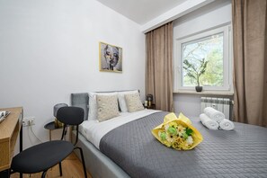 A hotel room with a bed, a desk, a chair, a window with curtains, and a painting on the wall. - Warsaw City Stay for families (Warszawa)
