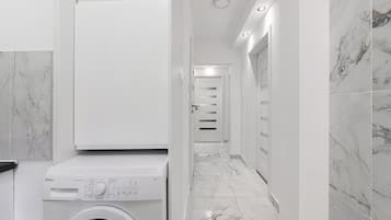 A modern laundry room with a front-loading washing machine, a hallway with white walls and marble flooring, and a door leading to another room.