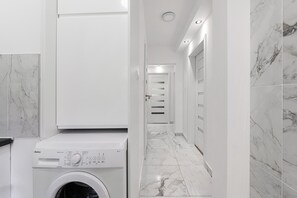 A modern laundry room with a front-loading washing machine, a hallway with white walls and marble flooring, and a door leading to another room. - Warsaw City Stay for families (Warszawa)