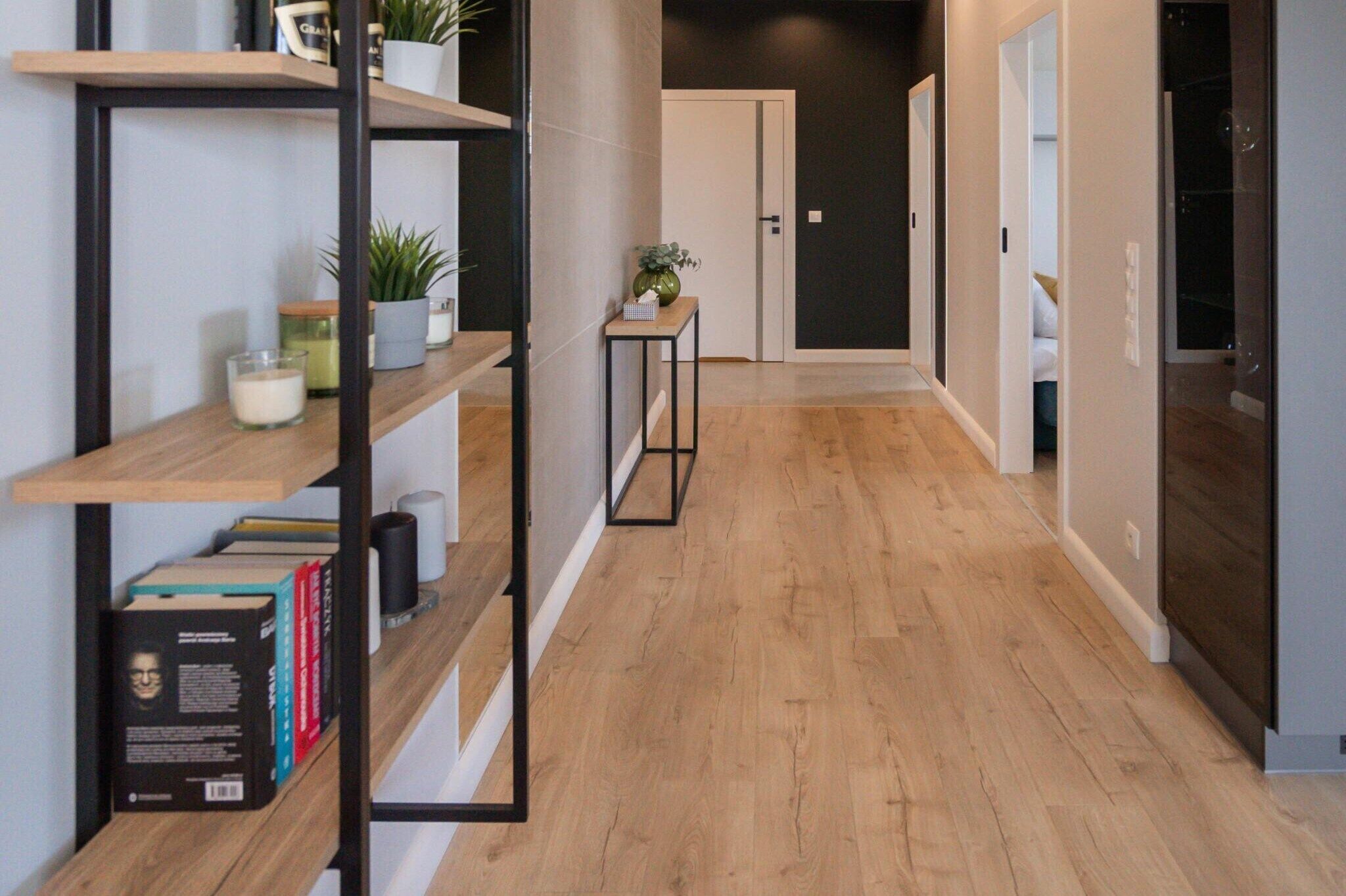 Un couloir doté d’un plancher en bois, une bibliothèque remplie de livres et d’objets décoratifs, une petite table avec une plante, et une porte au fond.