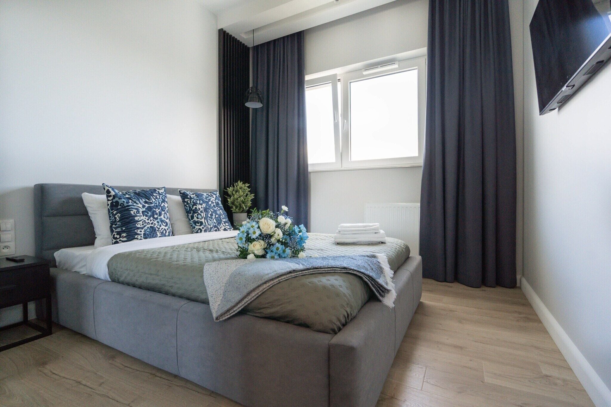 Un dormitorio moderno con un sofá cama gris, una mesita de noche, una ventana con cortinas y un televisor montado en la pared.