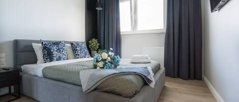 Un dormitorio moderno con un sofá cama gris, una mesita de noche, una ventana con cortinas y un televisor montado en la pared.