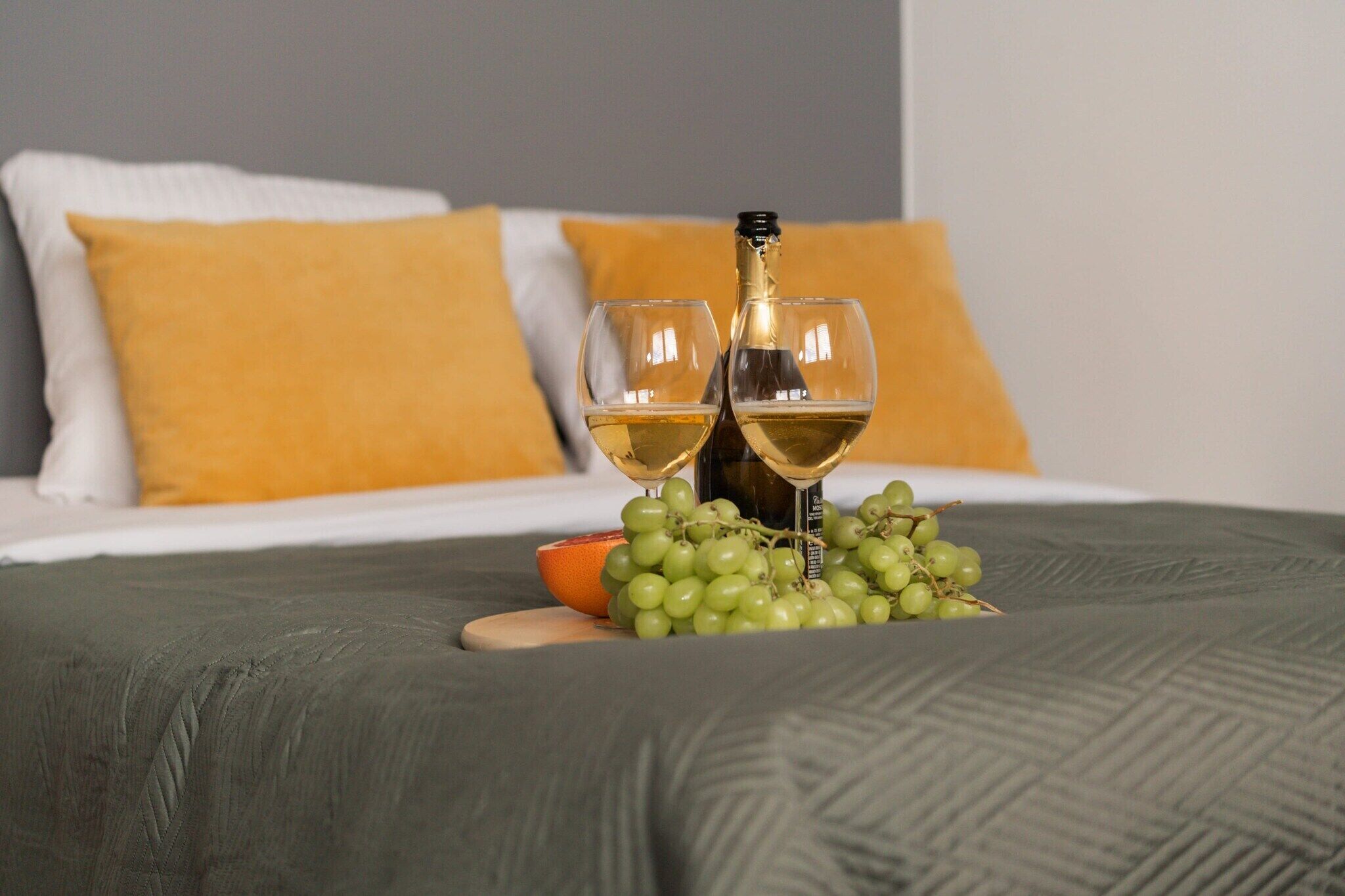 Una cama con un edredón verde, dos vasos de vino blanco, una botella de vino y un bol de uvas.
