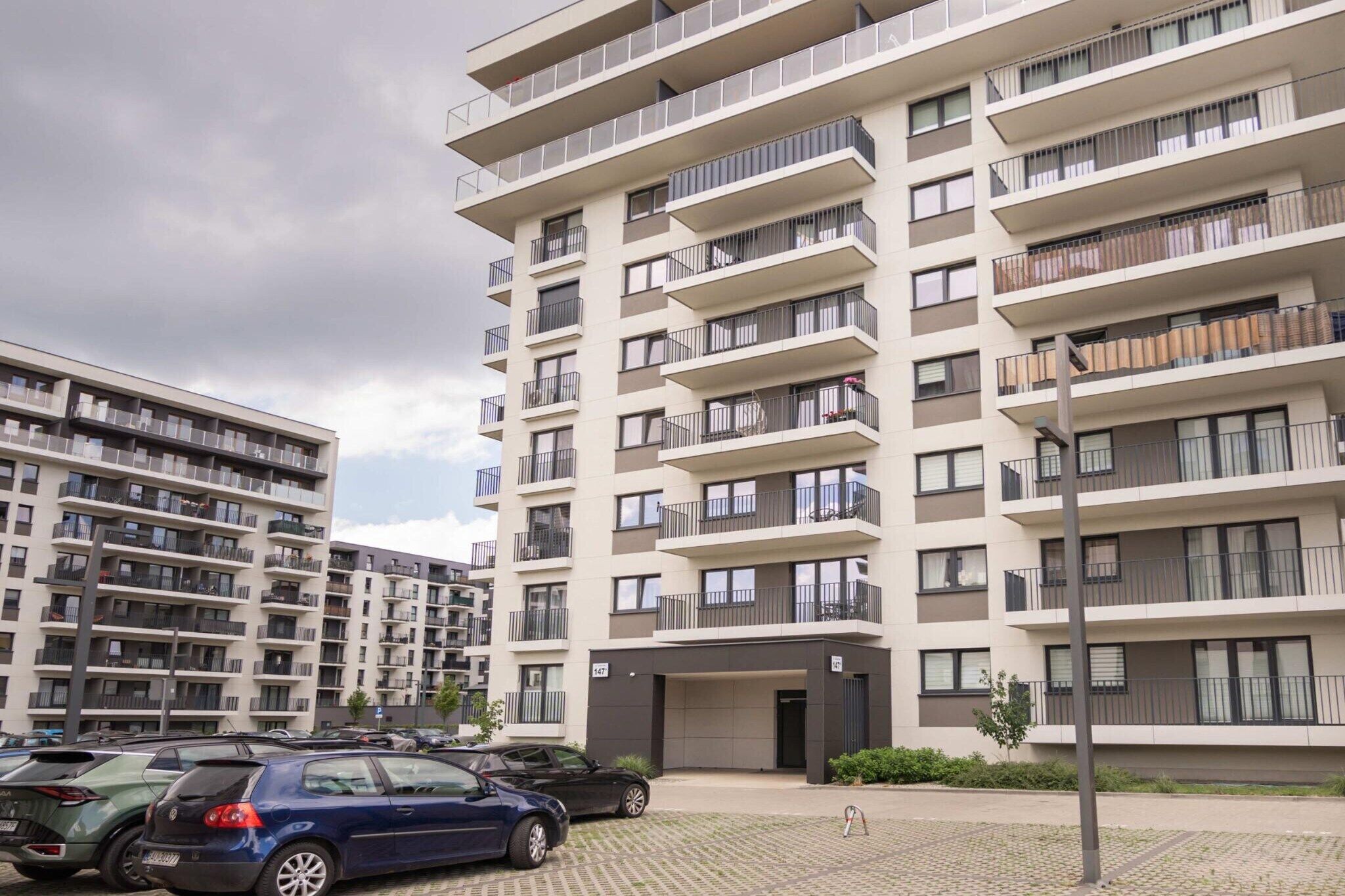 A multi-story residential building with balconies, a parking area, and a clear sky.