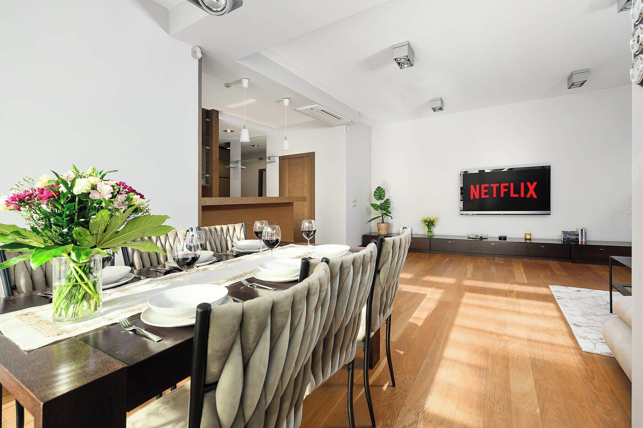 Un espace repas moderne avec une longue table dressée pour quatre personnes, un téléviseur affichant le logo de Netflix et une vue sur la cuisine.