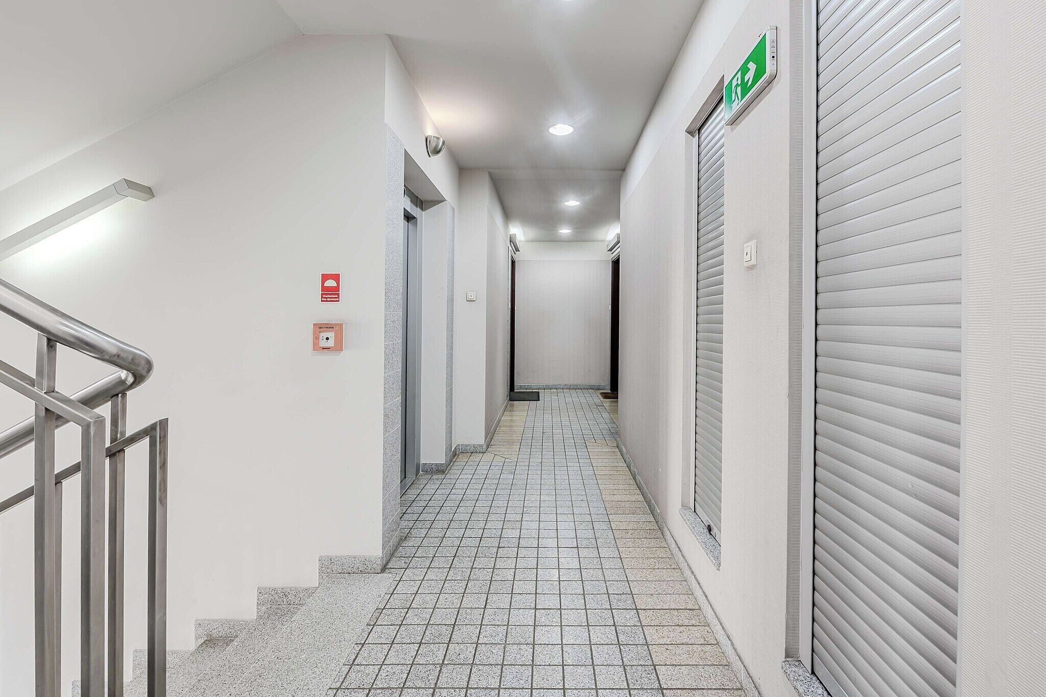 A corridor with a metal handrail, tiled floor, and closed shuttered doors on one side.