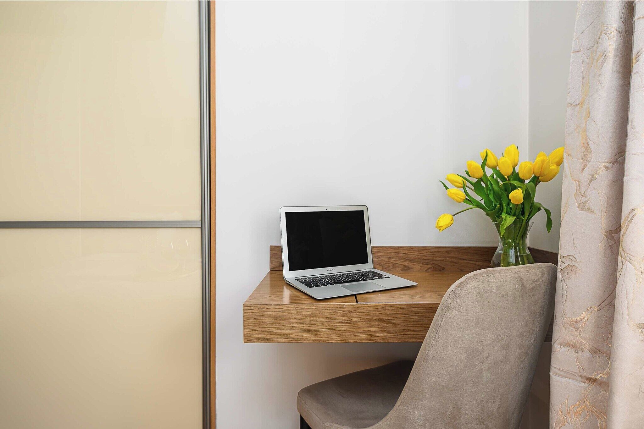 A laptop on a wooden desk, a chair, and a vase with yellow flowers.