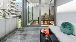 A balcony with a glass door leading to an outdoor dining area, a sofa with a cushion, and a table with a drink and fruit. - Parkside Flat with Private Garage (Łódź)