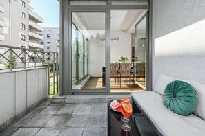 A balcony with a glass door leading to an outdoor dining area, a sofa with a cushion, and a table with a drink and fruit. - Parkside Flat with Private Garage (Łódź)