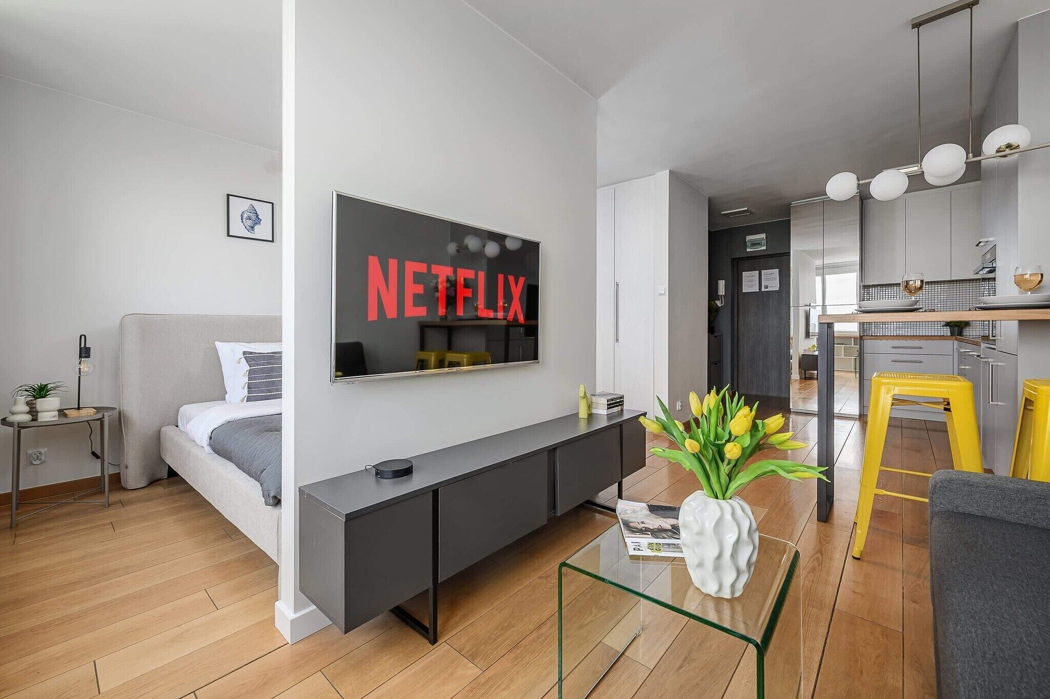 Een moderne woonkamer met een flatscreen televisie aan de muur, een bank, een salontafel, en een keuken in de achtergrond.