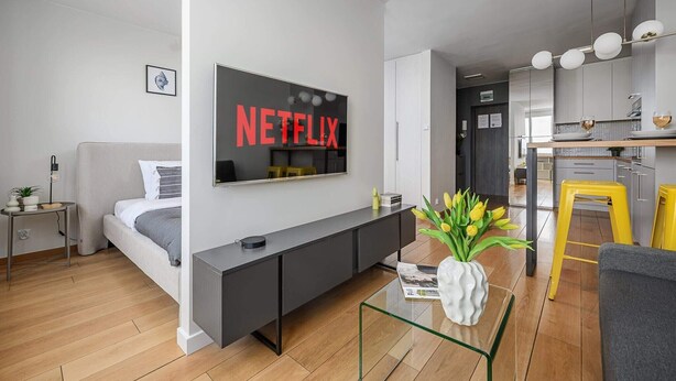 A modern living room with a flat-screen TV mounted on the wall, a sofa, a coffee table, and a kitchen area visible in the background. - Riverside Flat near Center (Warszawa)