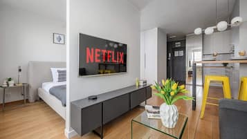 A modern living room with a flat-screen TV mounted on the wall, a sofa, a coffee table, and a kitchen area visible in the background.