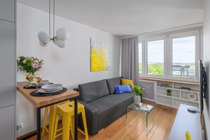Ein modernes Wohnzimmer mit grauem Sofa, einem Holzesstisch, gelben Hocker, einem Glastisch und Blick auf die Stadt durch große Fenster.