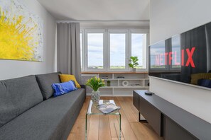 Ein modernes Wohnzimmer mit einem grauen Sofa, einem Couchtisch mit Zeitschriften, einem Fernseher mit dem Netflix-Logo und einem Fenster mit Blick auf den Himmel.