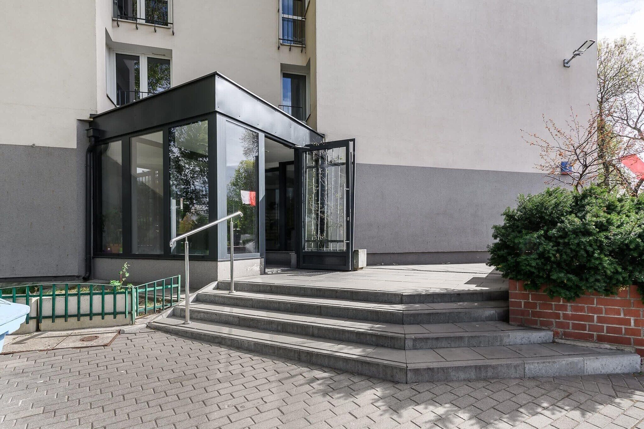 A building entrance with a glass door, steps leading up to it, and a small green fence.