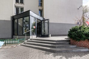 Ein Gebäudeeingang mit Glastür und Stufen, vor einer kleinen grünen Umzäunung.