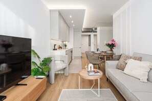 Een moderne woonkamer met een flatscreentelevisie, een grijze bank, een eettafel met stoelen, en een keuken in de achtergrond.