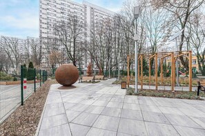 Ein gepflasterter Weg mit einer großen, kugelförmigen Skulptur, Bänken und einer Spielanlage.