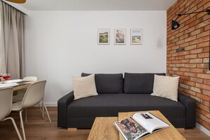 Una sala moderna con un sofá gris oscuro, dos almohadas blancas, una mesa de centro de madera con una revista, un escritorio blanco con una silla y una pared de ladrillo.