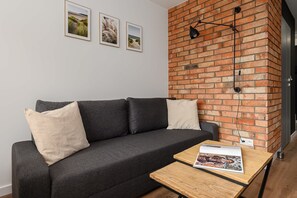 Ein modernes Wohnzimmer mit einem dunkelgrauen Sofa, zwei hellen Kissen, einem Couchtisch aus Holz mit einer Zeitschrift und einer Ziegelwand mit einer Pendelleuchte.