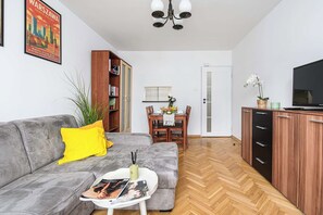 Un salon avec un canapé gris, une petite table basse, une bibliothèque, une table à manger avec des chaises, un téléviseur et un meuble en bois.