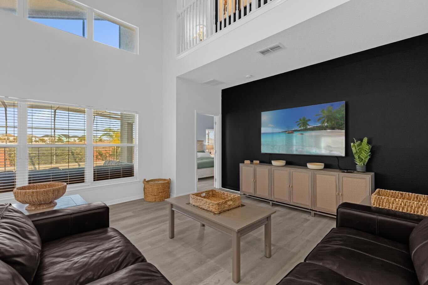 A spacious living room with a high ceiling, a large window with blinds, a black accent wall with a TV, and a view of the outdoors.