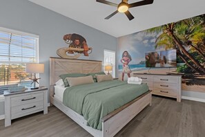 Ein Schlafzimmer mit einem Bett, Nachttischen, einem Deckenventilator, einem Fernseher und einem Wandgemälde mit einer Strandansicht.