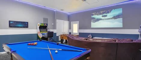 A pool table with balls, a foosball table, and a projection screen displaying a snowy landscape.