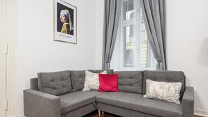 Ein graues Ecksofa mit vier Kissen, darüber ein gerahmtes Kunstwerk und ein Fenster mit grauen Vorhängen.