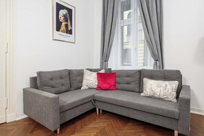 Ein graues Ecksofa mit vier Kissen, darüber ein gerahmtes Kunstwerk und ein Fenster mit grauen Vorhängen.