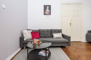 Ein Wohnzimmer mit einem grauen Sofa, zwei Kissen, einem Glastisch mit Zeitschriften, einem gerahmten Kunstwerk darüber und einer weißen Tür im Hintergrund.