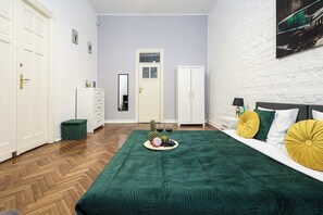 Une chambre avec un parquet en chevrons, un lit agrémenté d’oreillers verts et jaunes, une table de chevet, une commode, un miroir et une porte dotée d’un panneau en verre dépoli.
