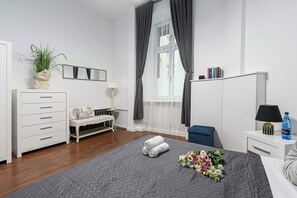 Ein modernes Schlafzimmer mit einer weißen Kommode, einem Sofa, einem Nachttisch, einer Lampe, einer Wasserflasche und einem Blumenstrauß.