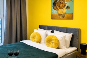 Ein Bett mit grauem Kopfteil, weißen und gelben Kissen sowie einer türkisblauen Bettdecke. Auf dem Bett stehen zwei Weingläser.