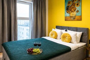A hotel room with a bed, a window with curtains, a painting on the wall, and a tray with grapes and wine glasses. - Modern Family Getaway in Łódź  CIty (Łódź)