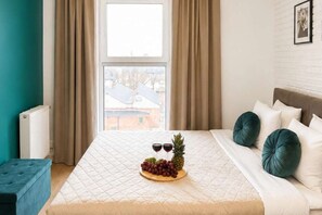 A bed with a quilted cover, a wooden tray with fruits and two glasses of red wine, and a view of a building through a window with curtains. - Modern Family Getaway in Łódź  CIty (Łódź)