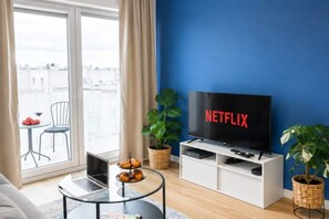 Ein modernes Wohnzimmer mit einem großen Flachbildfernseher, einem Laptop, einem kleinen runden Tisch mit Gebäck und Blick auf die Stadt durch ein großes Fenster.