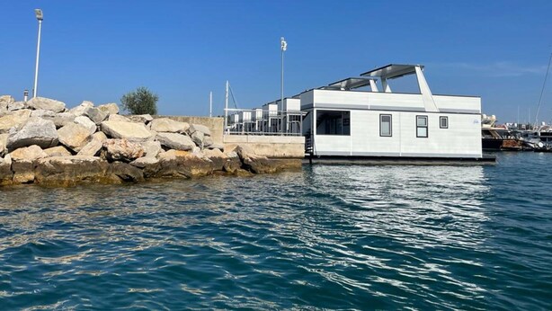 Ein Hausboot liegt in einer Marina vor einer felsigen Küste, der Himmel ist klar und blau.